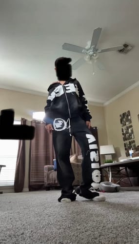 Reviewer models the black SEEK VISIONZ SET tracksuit with bold white graphics in a living room.