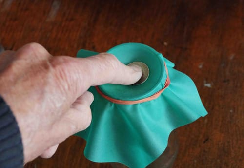 Reviewer performing the Gozinta coin trick, pushing a coin into the green rubber cover secured over a glass with a rubber band.