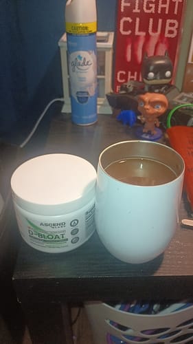 Customer's D-BLOAT powder container on a dark desk next to a prepared drink in a white cup.
