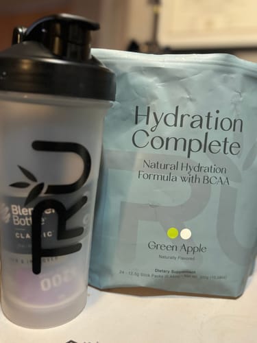 Reviewer's pouch of Hydration Complete Green Apple powder sitting on a desk next to a shaker bottle.