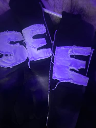 Close-up of a reviewer's black SEEK VISIONZ SET hoodie with large, distressed white lettering on the chest under purple light.