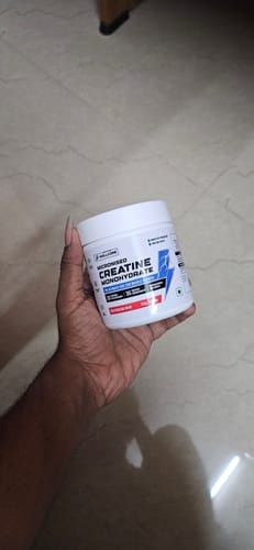 A reviewer holds up a white tub of Wellcore Pure Micronised Creatine Monohydrate against a tiled background.