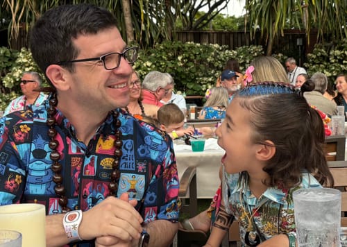 Customer wearing the Lilo & Stitch "Tiki-terrestrial" shirt with a lei, smiling at a child at an outdoor luau-style event.