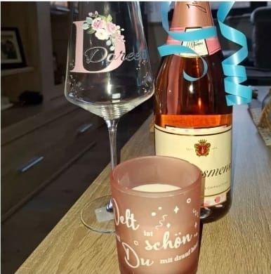 Rezensent zeigt ein Geschenkset mit einem personalisierten Weinglas, einer Flasche Wein und einer Tasse auf einem Tisch.