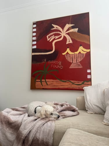 Customer's 'Paradise Found' framed art hanging on a wall above a couch where a small white dog is resting.