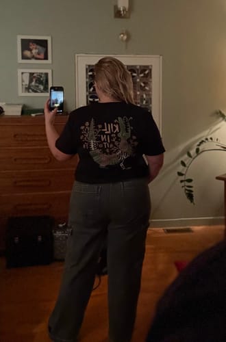 Reviewer taking a mirror selfie to show the back graphic and fit of the black Killin It T-Shirt.