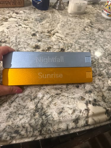 Customer holding two metal Weekly AM-PM Vitamin Cases, a blue 'Nightfall' case stacked on a gold 'Sunrise' case over a counter.