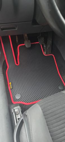 Recenzent pokazuje czarny dywanik samochodowy z czerwonym obszyciem, dopasowany po stronie kierowcy.