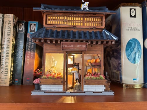 Reviewer's completed Ikseon-dong Hanok Bakery kit displayed on a bookshelf, illuminated and with a custom cat on the roof.