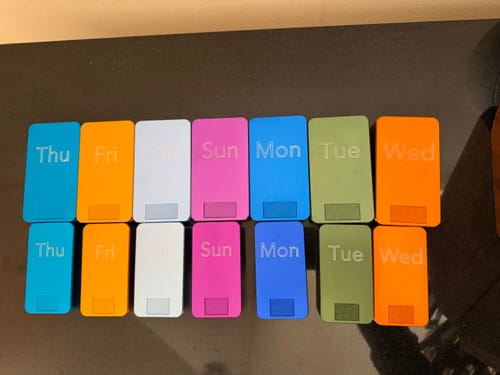 Customer's collection of 14 colorful metal pill cases organized by day of the week on a dark surface.