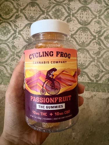 Customer holds a clear jar of Cycling Frog Passionfruit THC Gummies, showing the product label and the orange gummies inside.