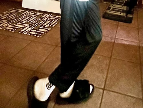 Customer wearing black furry DracoSlides with white socks and black pants on a tiled floor.