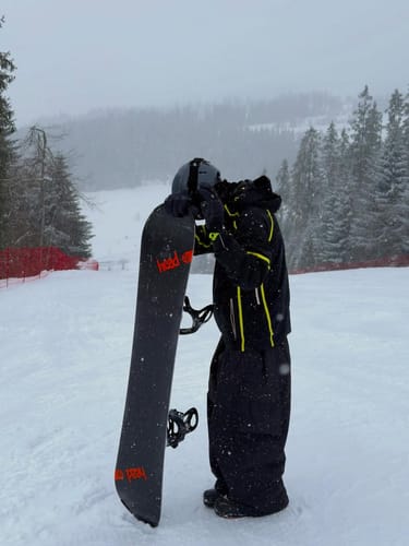 Reviewer wearing black baggy snow pants while holding a snowboard on a snowy slope.