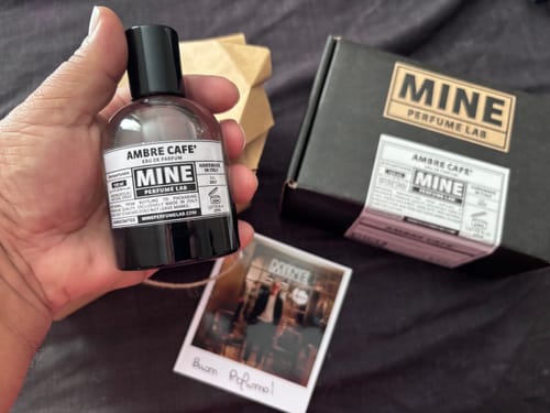 Un cliente tiene in mano la boccetta di profumo AMBRE CAFE' con la sua scatola nera e una cartolina sullo sfondo.