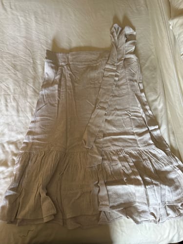 Una clienta muestra su Vestido Agatha color arena extendido sobre una cama, destacando su lazo y su bajo con volantes.