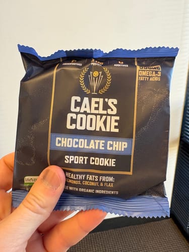 A reviewer's hand holding the dark packaging of a Cael's Cookie Chocolate Chip sport cookie.