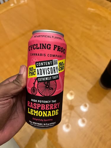 Customer's hand holding a pink can of 'Extremely Tasty' THC Raspberry Lemonade over a wooden table.