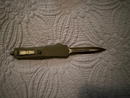 Customer's Black Ghost OTF knife with its dagger-style blade extended, displayed on a gray quilted surface.
