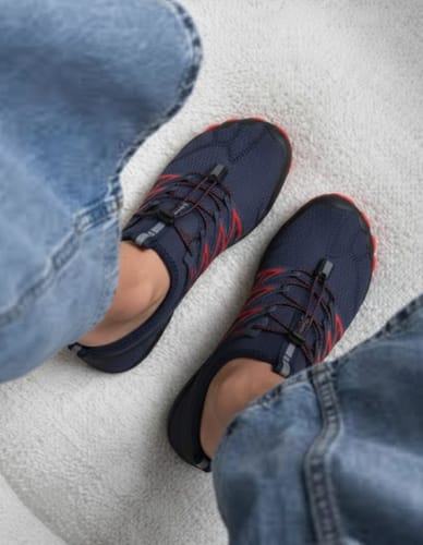 Customer's feet viewed from above, wearing dark blue and red BareEase shoes with jeans on a light-colored rug.