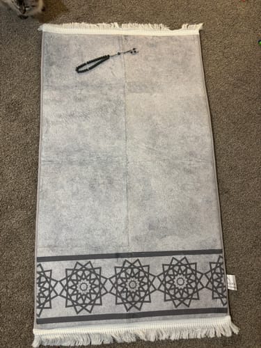 Customer's grey prayer mat laid on a carpet, showing its geometric border. The gifted tasbih is also visible on top.