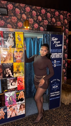 Customer wearing the brown Butter Turtleneck Contour Bodysuit with a chain belt, posing by a vintage photo booth.