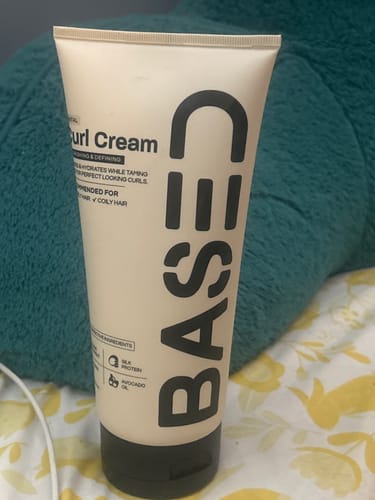 Customer's beige tube of Based Curl Cream resting against a plush teal pillow.