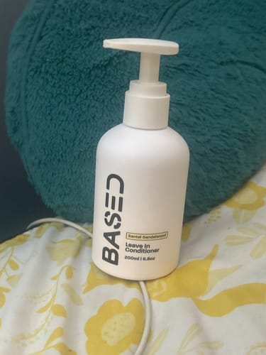 Customer's white pump bottle of Based Bodyworks Leave-In Conditioner resting on a bed.