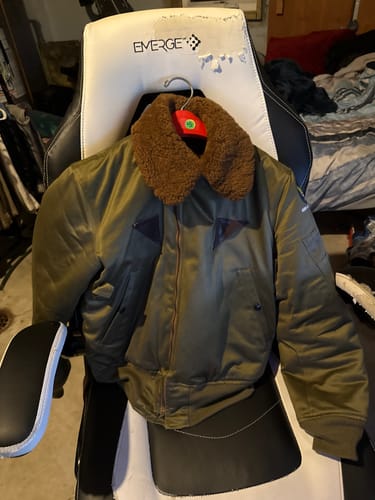 Reviewer's olive green USAAF Type B-15A Flight Jacket with a brown sherpa collar, hanging on a chair.