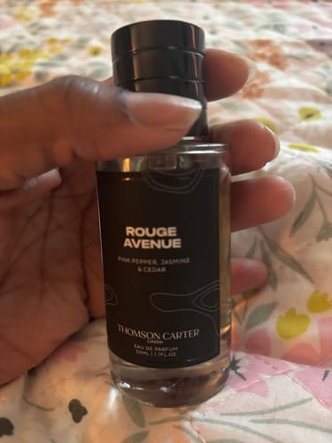 Customer's hand holding the clear glass bottle of Rouge Avenue Eau de Parfum, showing the black label.