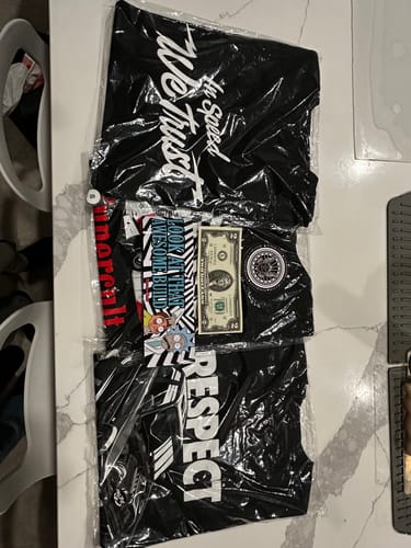 Reviewer's contents from the TCG92 MYSTERY CASH BOX, including three T-shirts, stickers, a patch, and a dollar bill.