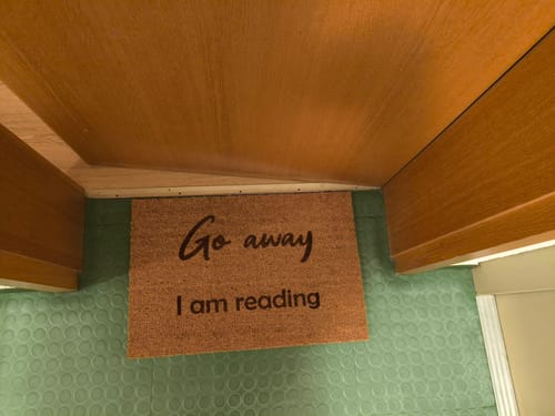 Rezensent zeigt die personalisierte Kokos-Fußmatte mit dem Spruch „Go away I am reading“ vor einer Tür.