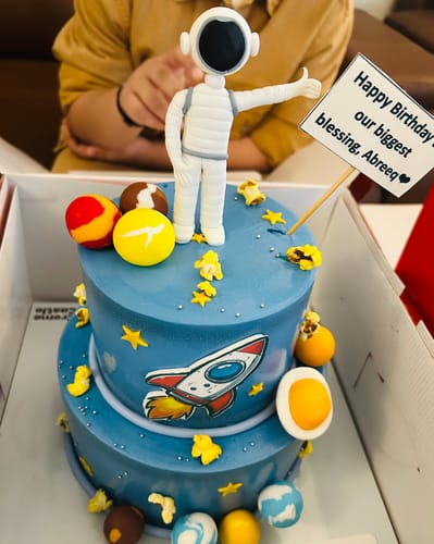 Customer's two-tier blue Universe Theme Cake with an astronaut topper and planets, displayed in its open box.