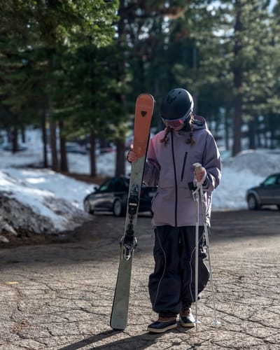 Reviewer wearing the lavender snow jacket with a baggy fit, holding skis in a snowy area.