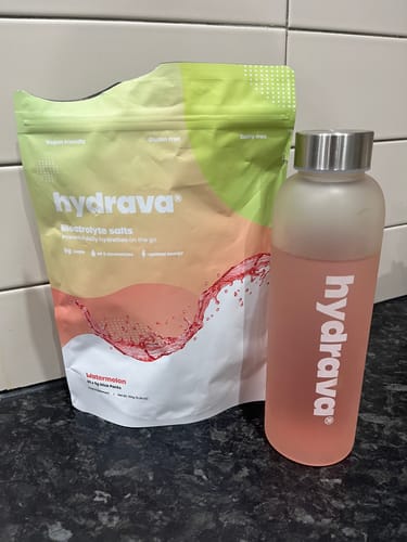 Customer's Frosted Hydrava Bottle with a pink drink, next to a pouch of Hydrava watermelon electrolyte salts on a counter.
