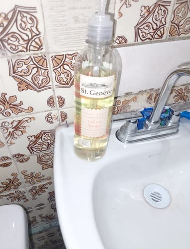 Cliente muestra el jabón líquido St. Geneve en su botella con dosificador sobre un lavabo.