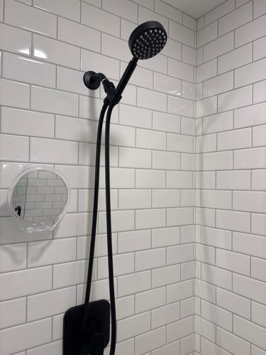 Reviewer's black handheld shower head set installed against a white subway tile wall in a shower.
