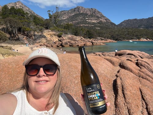 Reviewer holding a bottle of Little Ripples Bubbles wine in a selfie at a sunny, rocky bay.