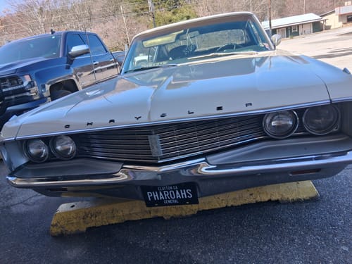 Customer's black metal license plate with the word 'PHARAOHS' mounted on the front bumper of a classic white car.