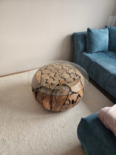 Kunde zeigt den runden Wohnzimmertisch XILON aus Teakholz mit Glasplatte, der auf einem beigen Teppich neben einem Sofa steht.