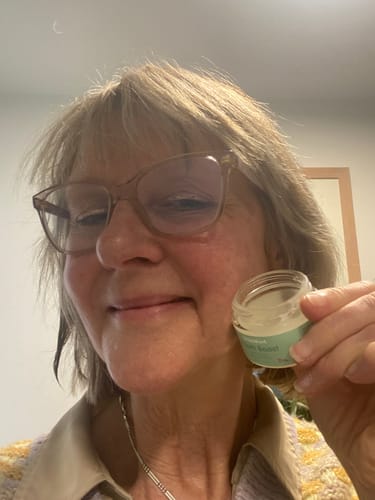 Reviewer smiling while holding an open glass jar of Youth Boost serum next to her cheek.