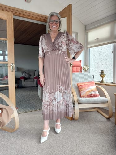 Reviewer smiling while wearing the Blaise Taupe Floral Print Wrap Dress, showing its pleated skirt and angel sleeves in a sunroom.
