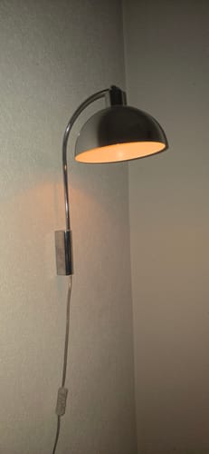 Klant toont de ingeschakelde Nordlux Ellen wandlamp met chromen afwerking, gemonteerd in een hoek.