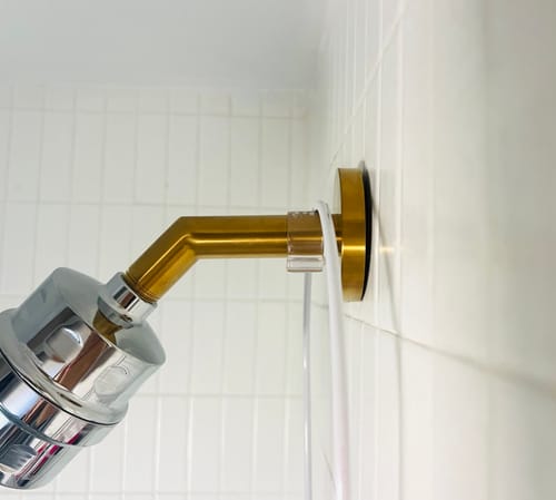 Close-up of the customer's gold 7 Inch Shower Arm and Flange installed on a white tiled wall with a chrome filter attached.