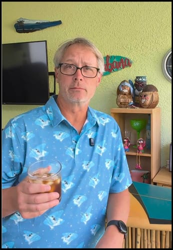 Reviewer in a tiki-themed setting wearing the blue patterned "Mid-Century Mando" All-Day Polo while holding a drink.