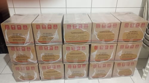 Cliente mostra várias caixas de papelão lacradas da Cesta Básica Cedro empilhadas em um cômodo.