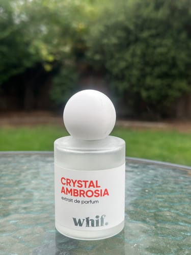 Reviewer's frosted bottle of Crystal Ambrosia perfume with a round cap on an outdoor table.