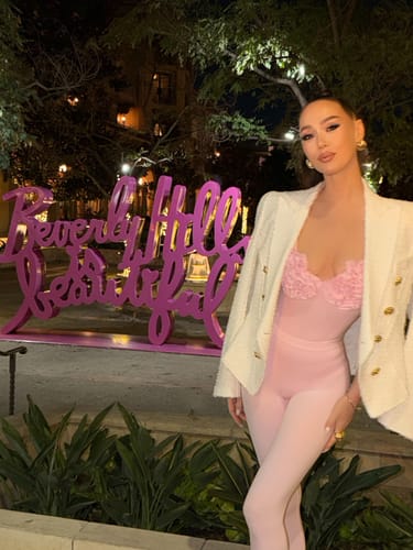 Reviewer styling the pink Naked Rose Tank Bodysuit with a white jacket in front of a neon sign.