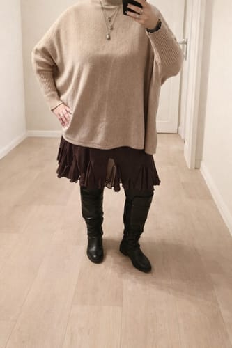Kundin trägt den beigefarbenen Oversize Cozy Pullover über einem dunklen Rock und Stiefeln.