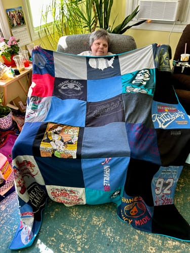 A customer sits in a sunlit room, holding up their large Premium T Shirt Quilt made of colorful shirt squares.