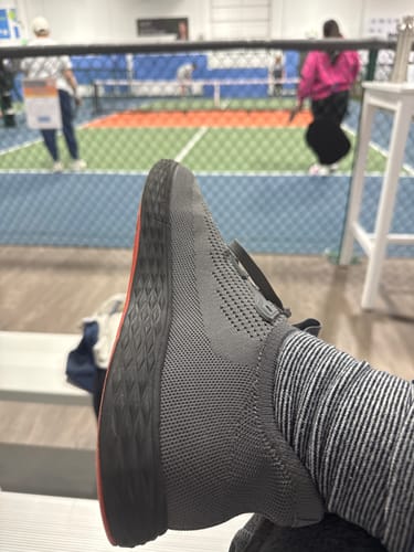 Reviewer wearing the grey Stride Pickleball Shoes while resting on a bench next to an indoor court.